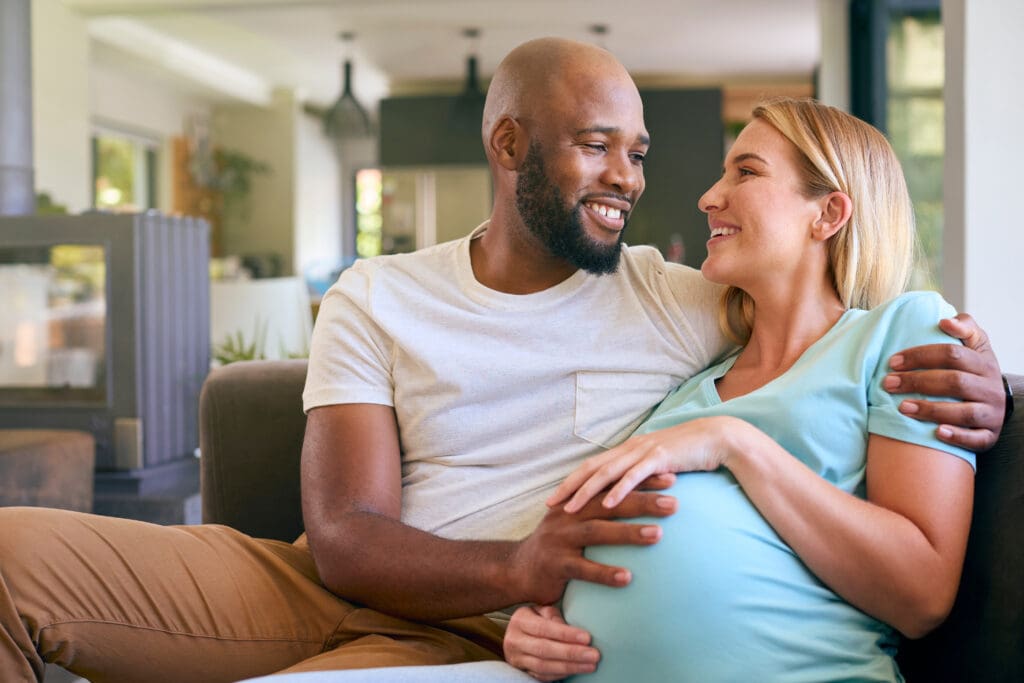 Loving Multi-Racial Couple With Pregnant Woman On Sofa At Home With Multi-Generation Family Behind