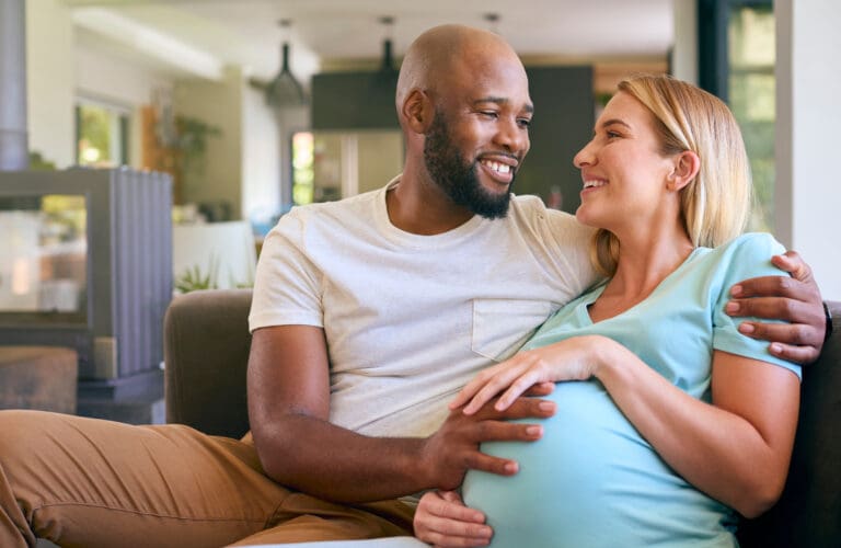 Loving Multi-Racial Couple With Pregnant Woman On Sofa At Home With Multi-Generation Family Behind
